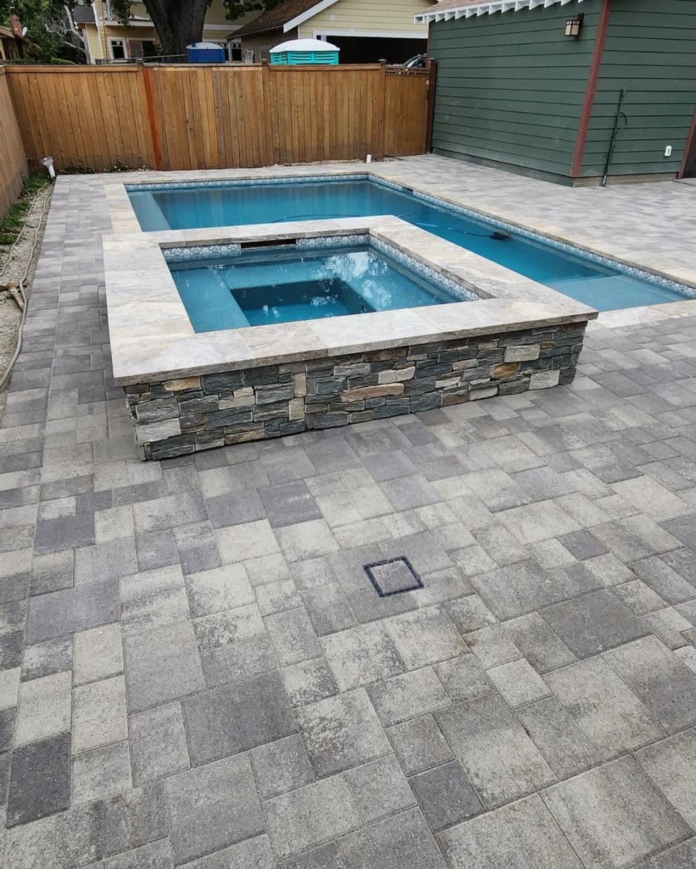 Modern backyard with a rectangular pool and spa, surrounded by stone coping and paved patio.