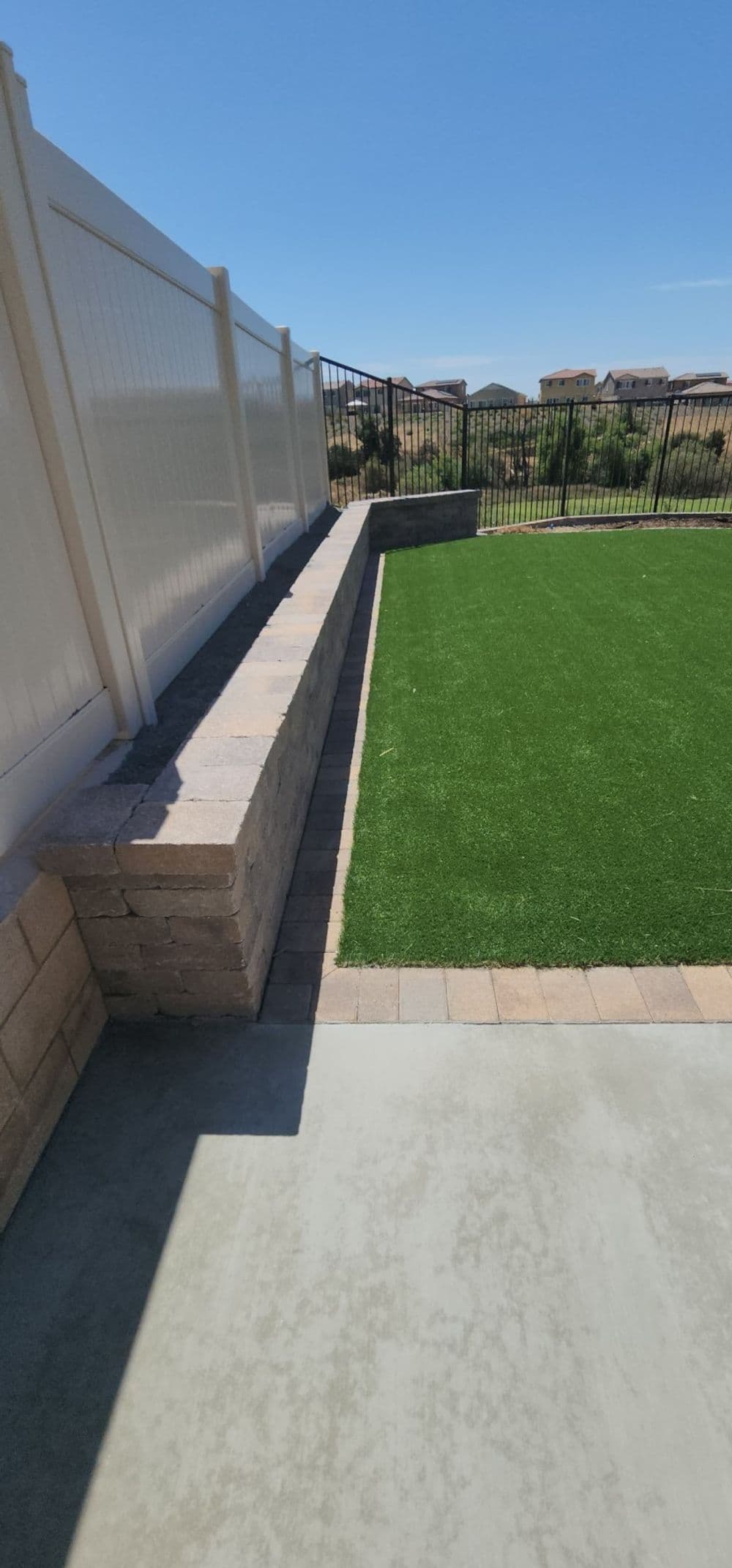 Lush green artificial turf next to a stone border and white fence in a sunny backyard.