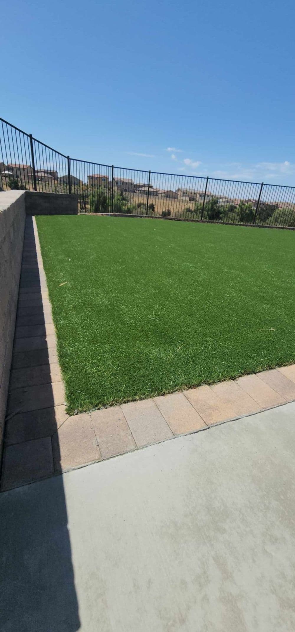 Lush green artificial grass lawn with a stone border and fencing against a clear blue sky.