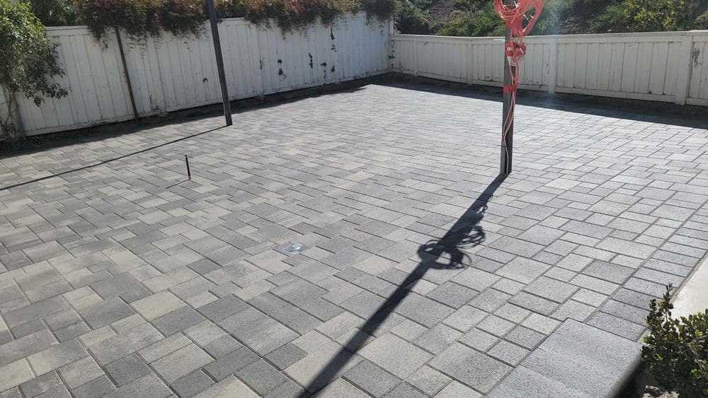Paved backyard patio featuring gray interlocking stones and a shadow from a pole.