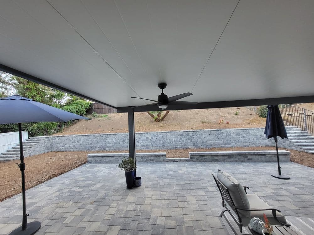 Covered patio area with gray pavers, ceiling fan, and umbrellas overlooking a landscaped yard.