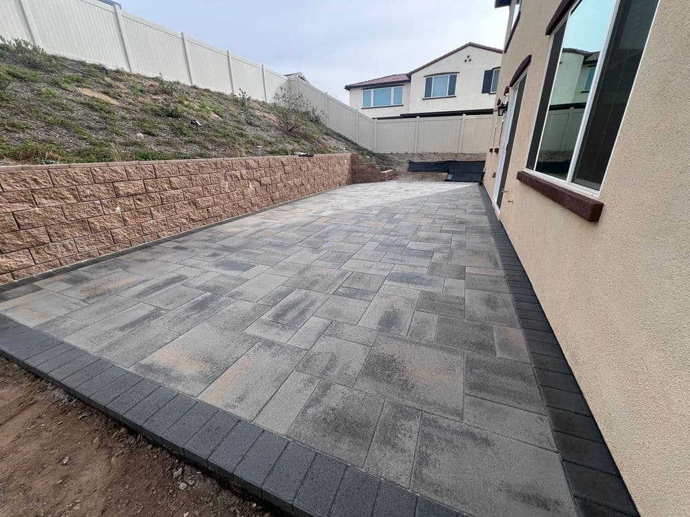 Paved patio area with gray stones and a retaining wall beside a house.