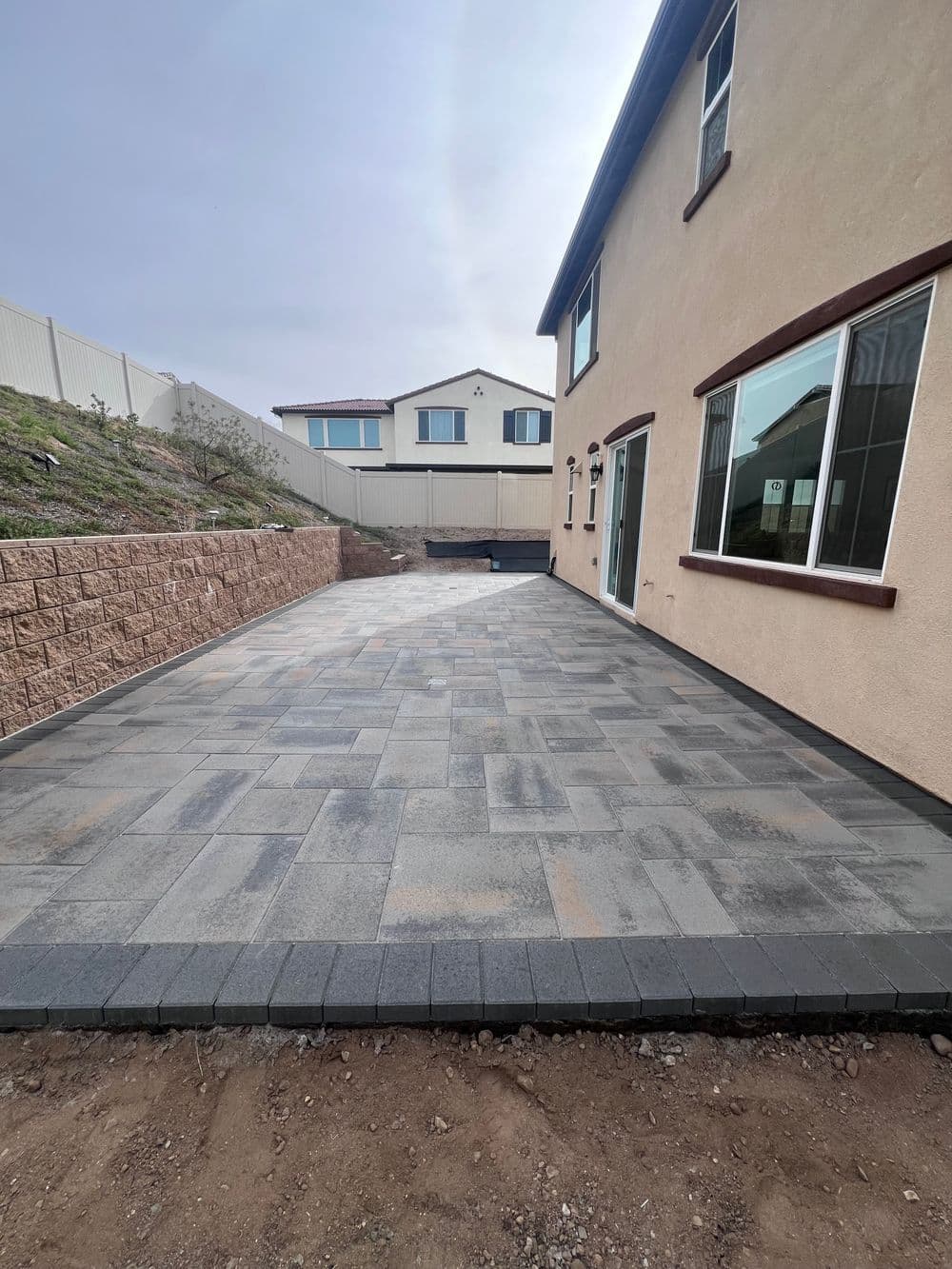Newly paved outdoor patio with gray pavers beside a residential home and landscaped area.