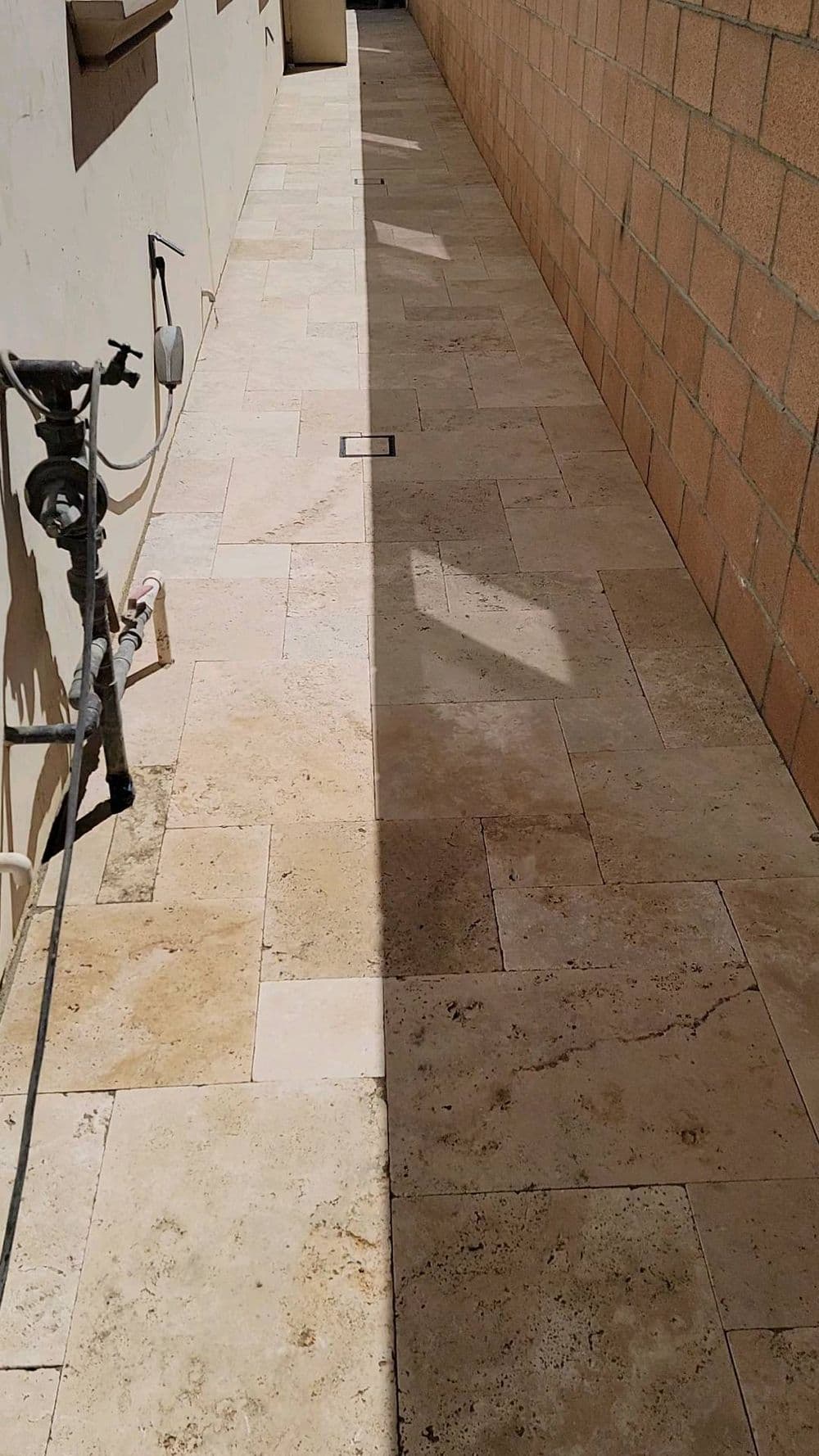 Narrow stone-tiled walkway with shadows and utility pipes along a brick wall.