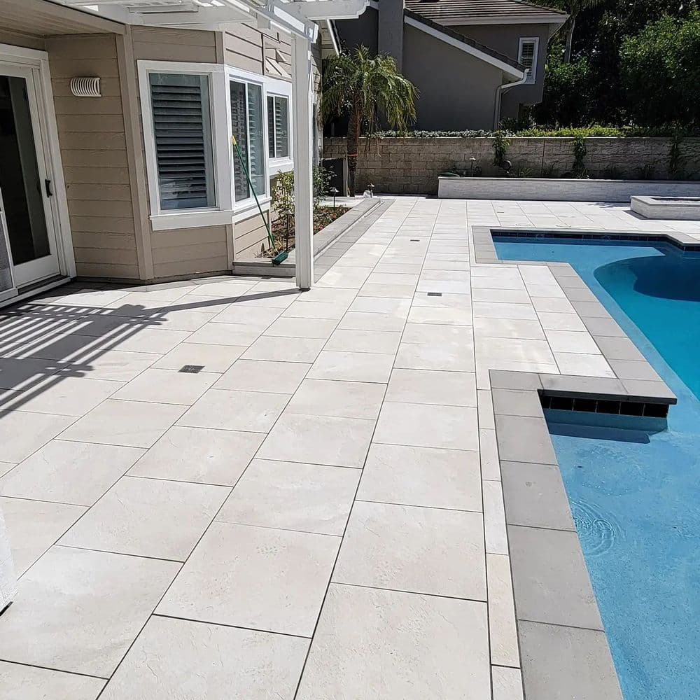 Modern patio with light-colored tiles, surrounded by a pool and landscaped greenery.